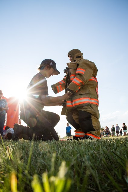 A person assists another in putting on a firefighter suit outdoors, with spectators in the background and sunlight shining.