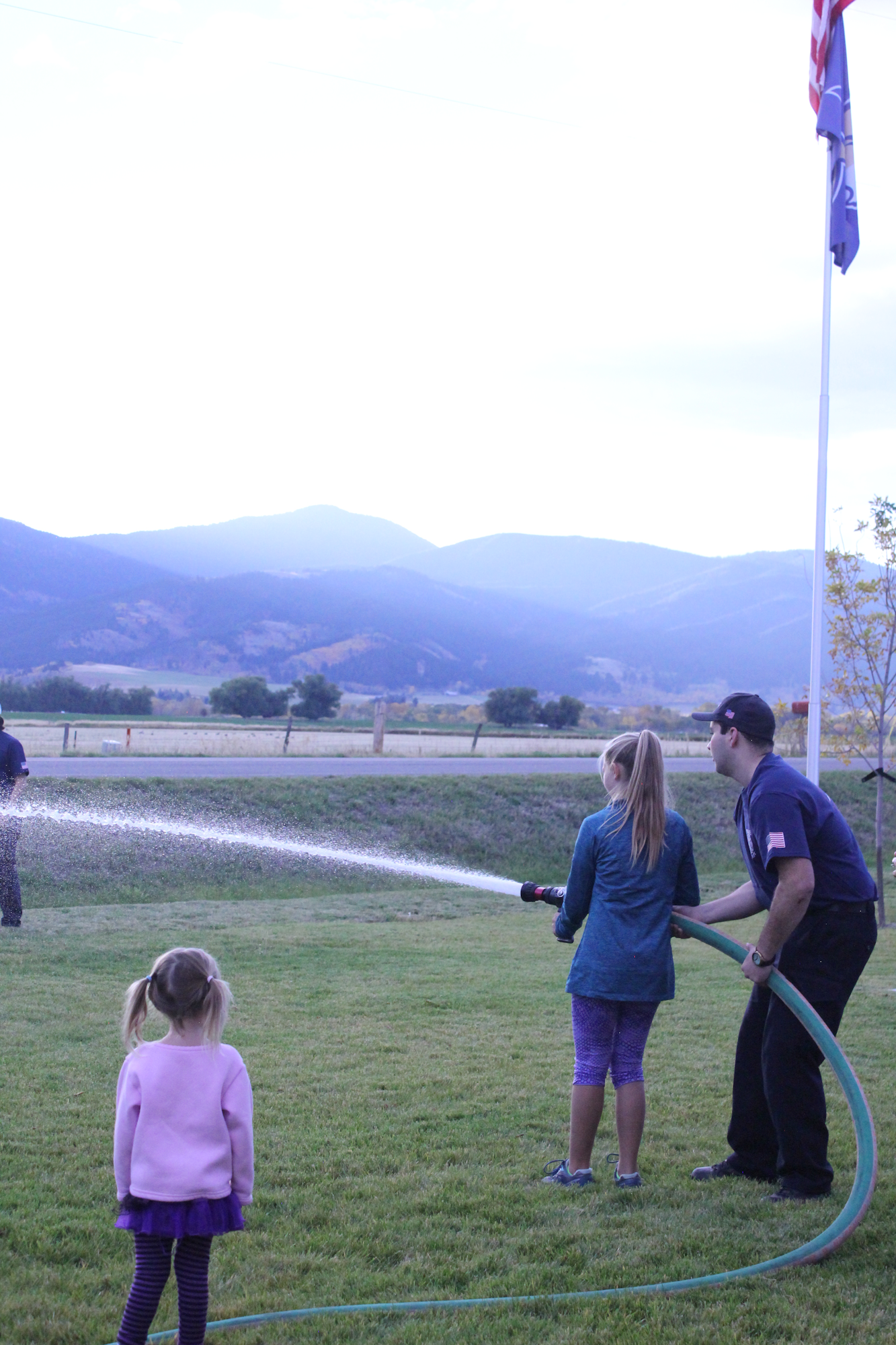 A young girl watches as two people spray water from a hose in a grassy area against a backdrop of mountains and flags.