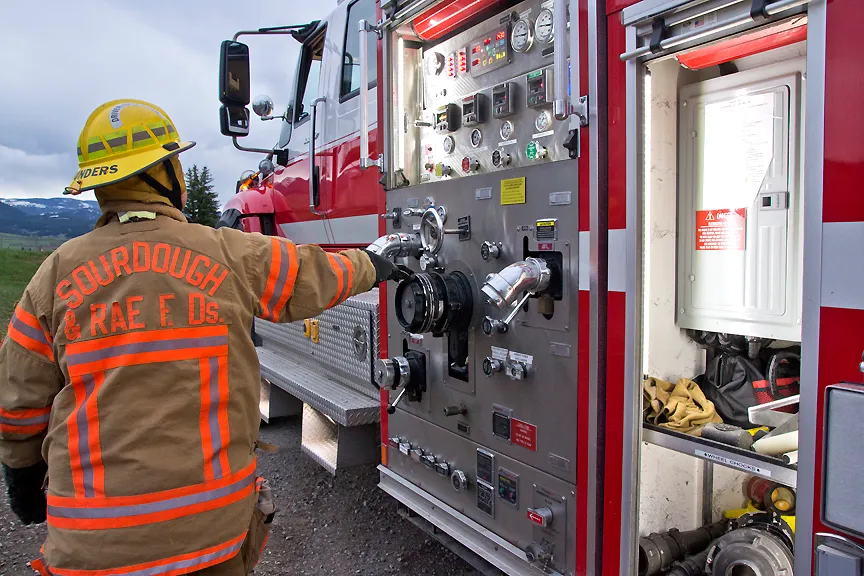 A firefighter in a yellow and red uniform operates a control panel on a fire truck, surrounded by mountains and clouds.