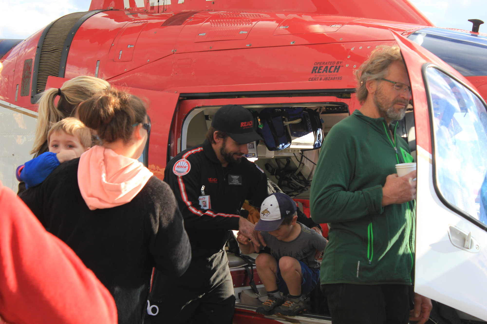 A crowd gathers around a red helicopter, with a medical team member assisting children and adults during an event.