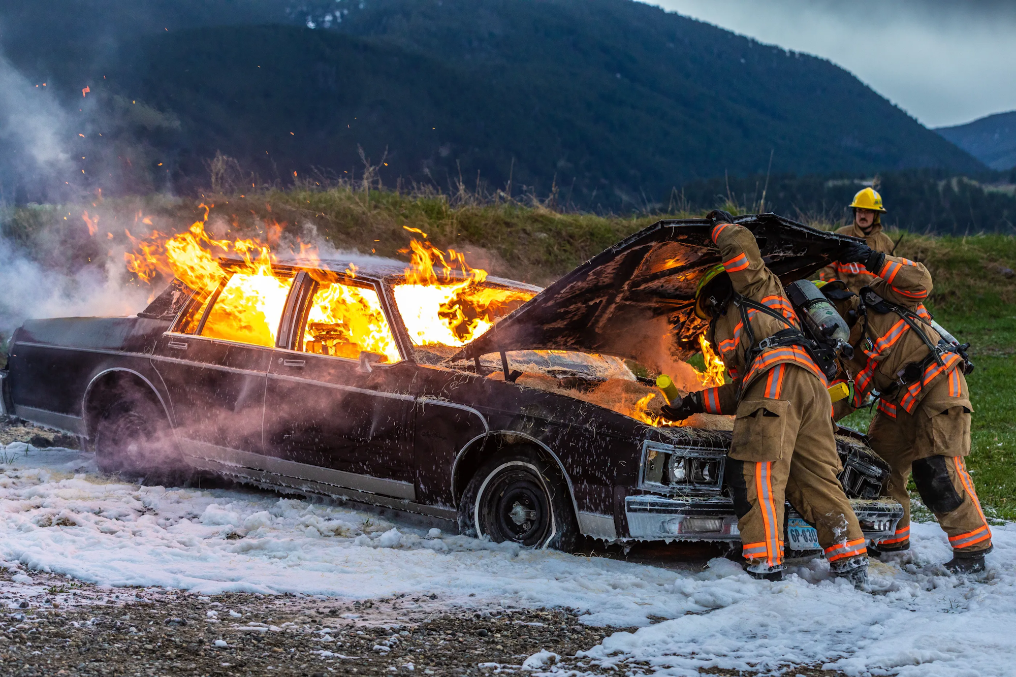 Firefighters are combating a fire in a car, using foam while smoke and flames are visible against a scenic backdrop.