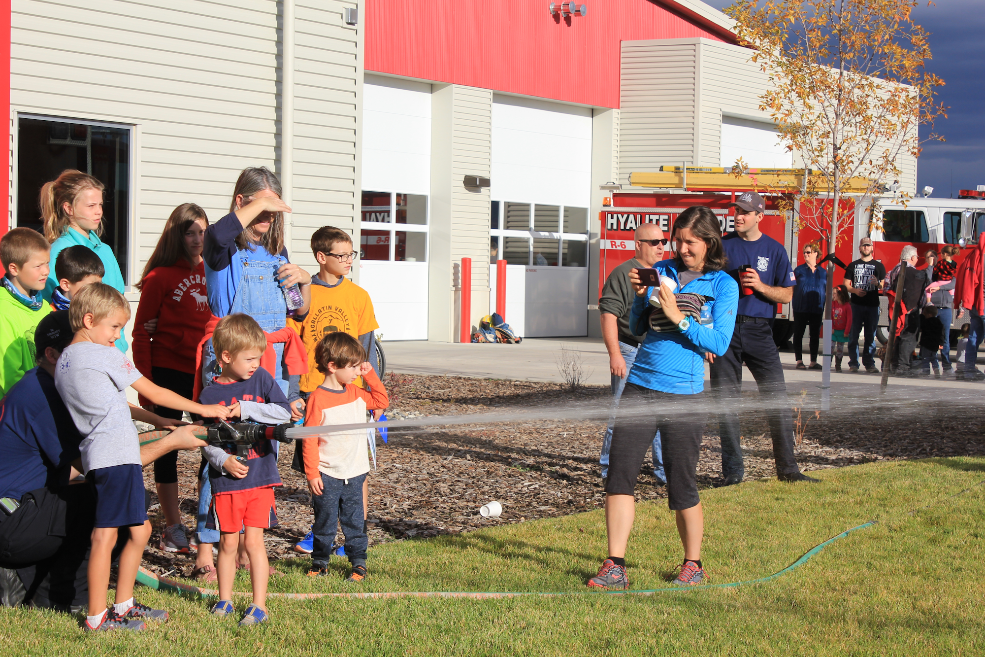 A group of children and adults watch as a child demonstrates using a fire hose, with a fire truck in the background.