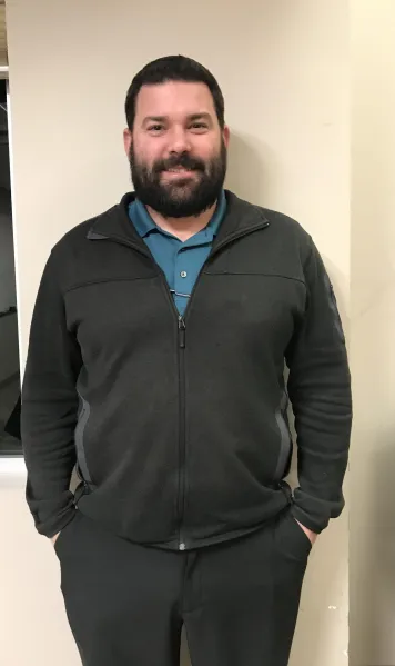 A man with a beard is standing casually, wearing a black zip-up jacket over a blue shirt, with hands in his pockets.