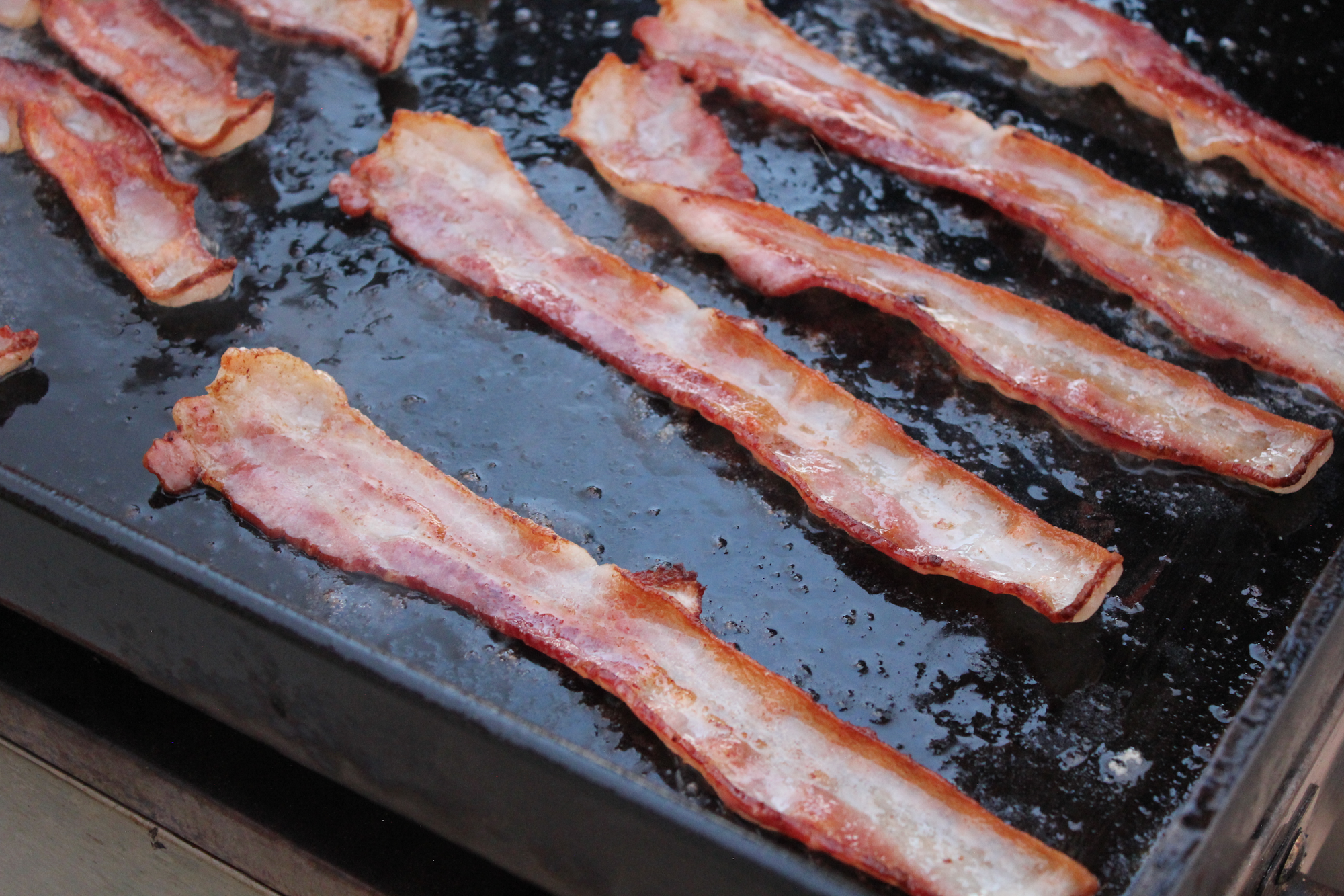 The image shows strips of bacon cooking on a black frying surface, sizzling with grease.