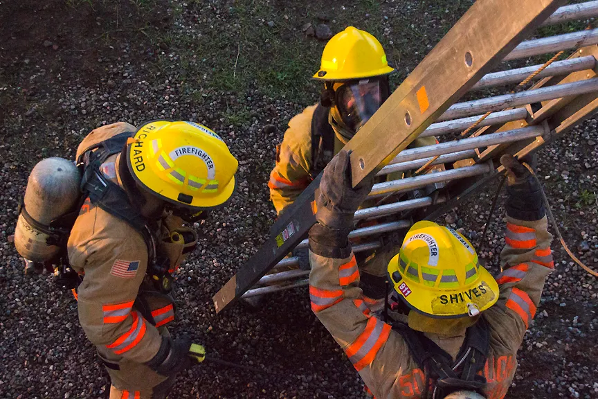 Three firefighters in gear are working together to lift a ladder against a gravel background, showcasing teamwork and safety.