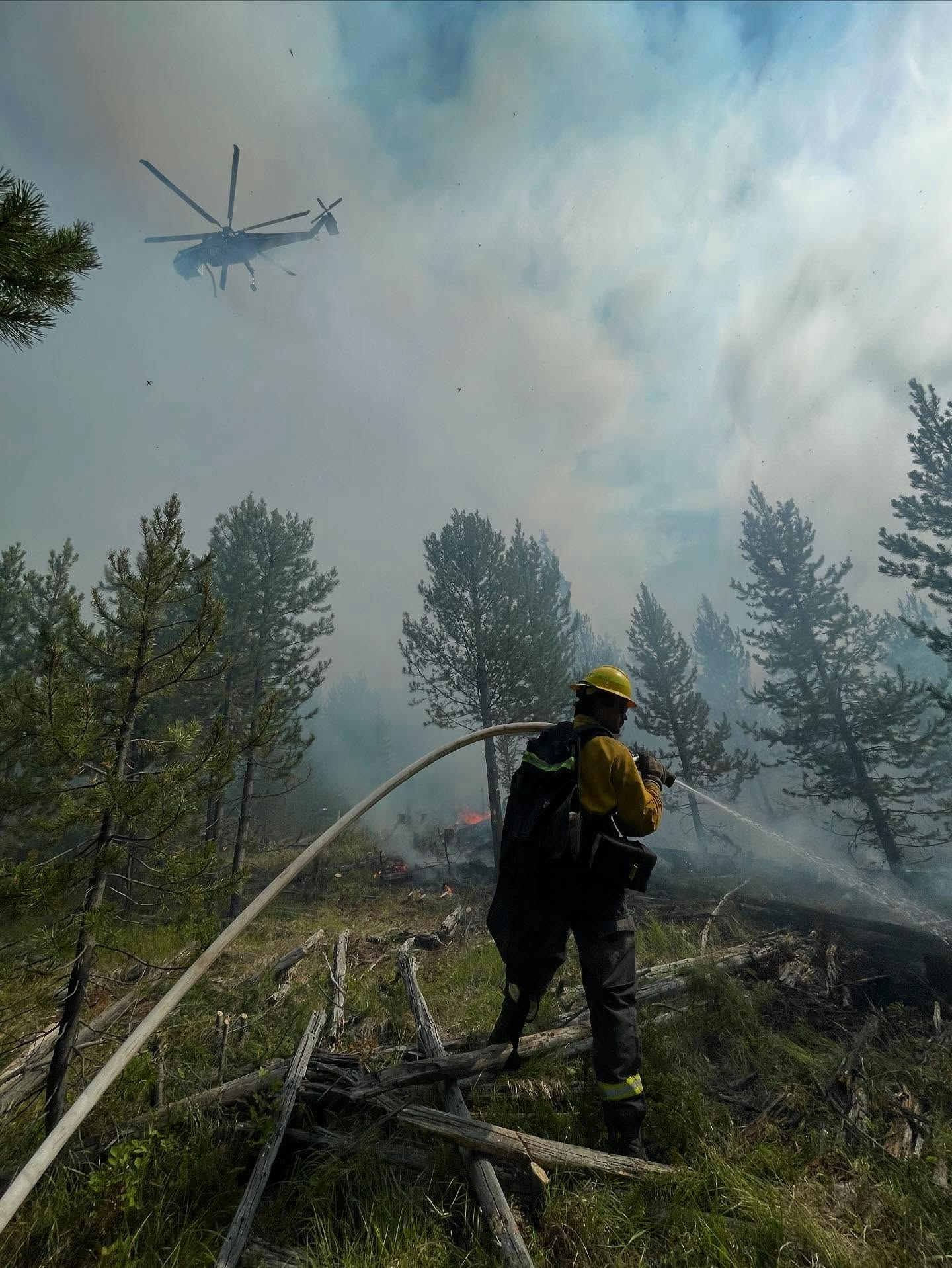 A firefighter uses a hose to combat a wildfire, with a helicopter above and smoke filling the air among the trees.