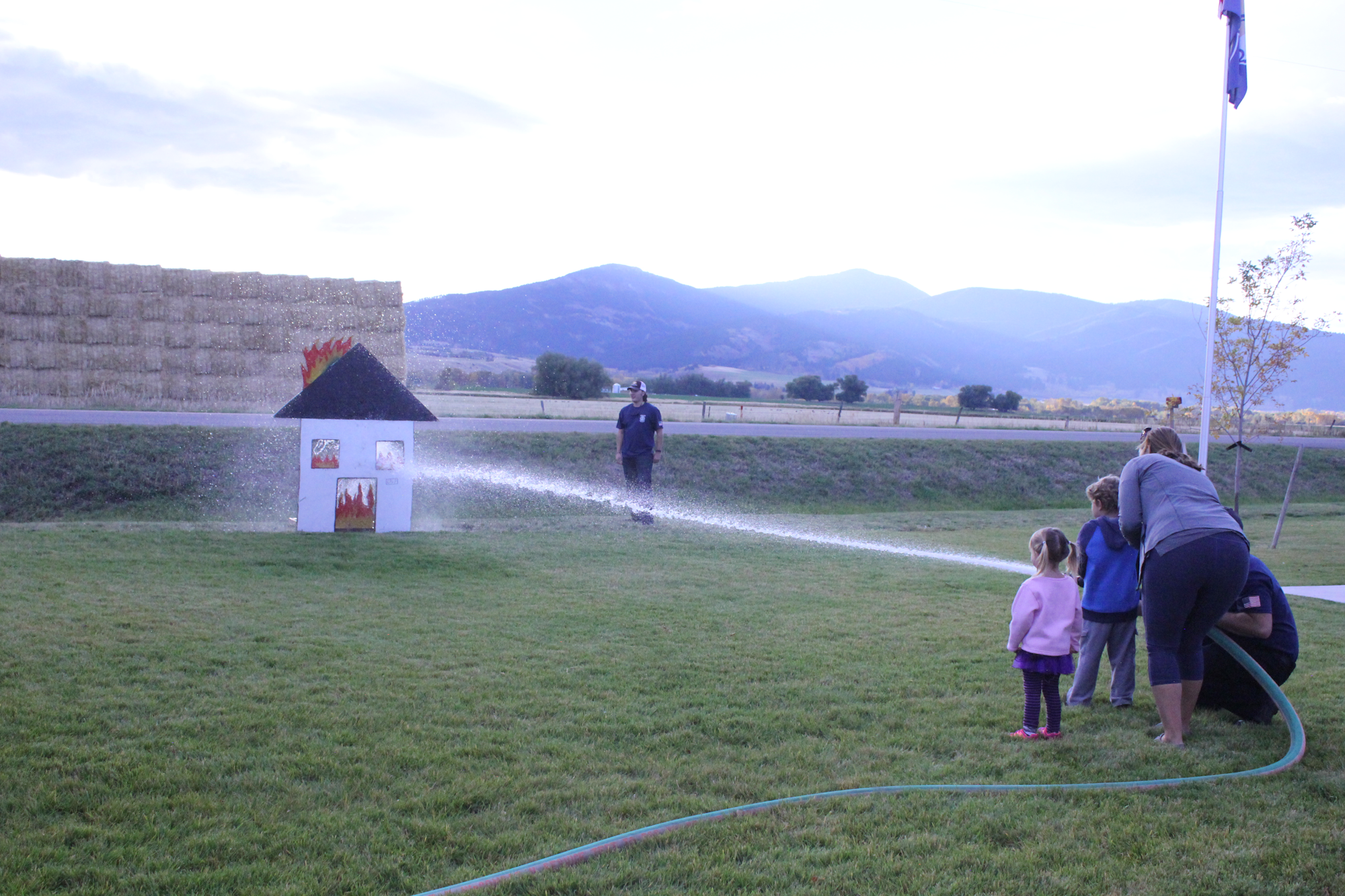 A firefighter demonstrates using a hose to put out a simulated fire on a small structure while kids watch attentively.