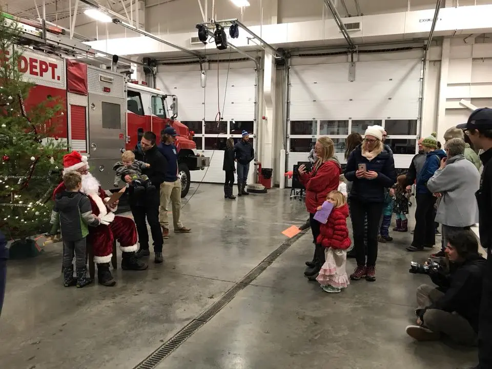 A festive scene at a fire station with Santa, children, a Christmas tree, and families enjoying holiday activities.