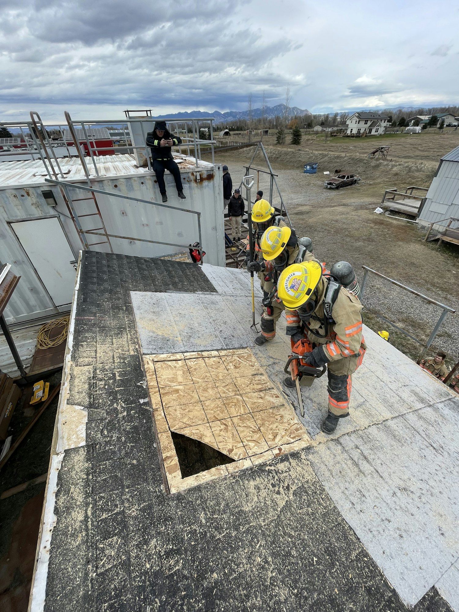 Firefighters are working on a roof, cutting through material, with a person observing nearby. Mountains and structures are in the background.
