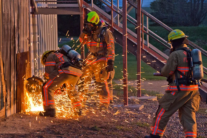 Firefighters use equipment to cut through a metal surface, creating sparks, with one firefighter standing watch.