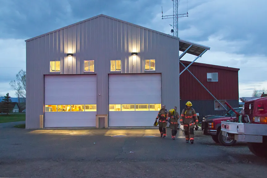 Firefighters in gear walking toward a lit fire station with large garage doors, under a cloudy evening sky.