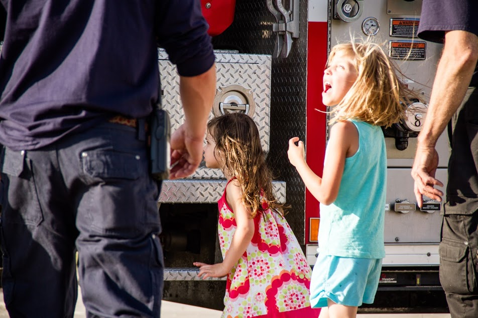 Two excited young girls are near a fire truck, interacting with adults, likely enjoying a community event.