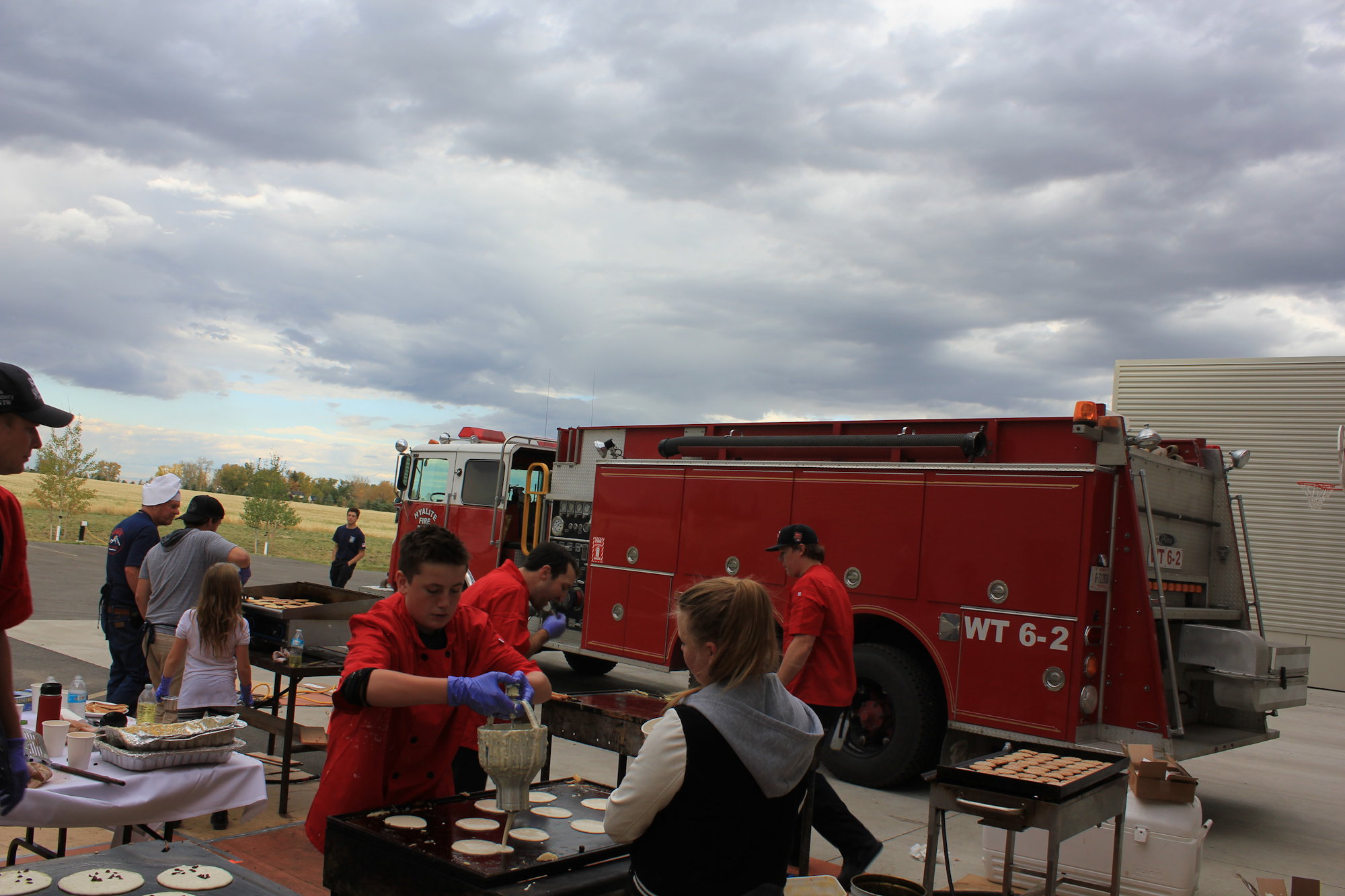 People are cooking pancakes outdoors, with a fire truck in the background under a cloudy sky.