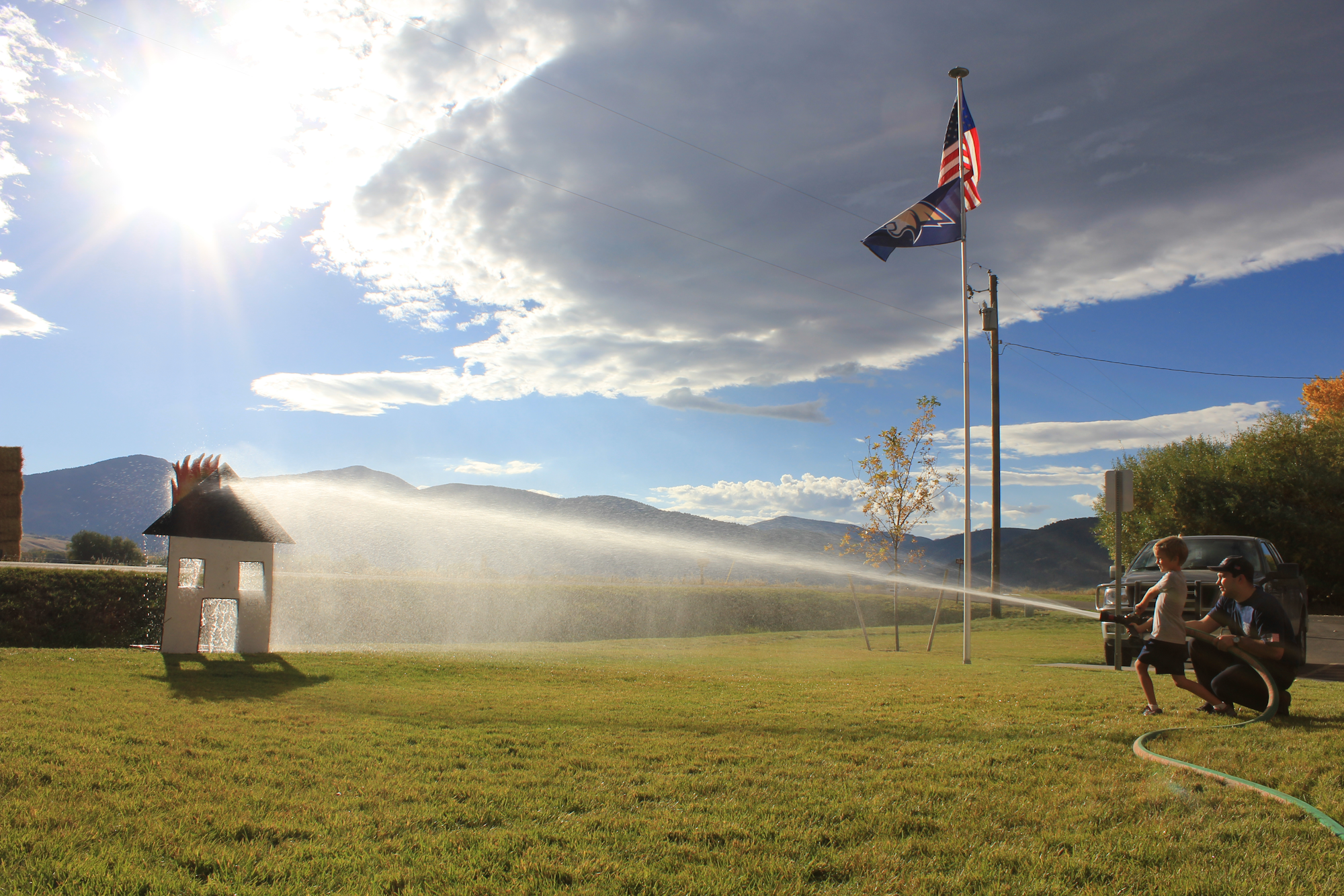 A child and an adult spray water from a hose towards a small playhouse, with mountains and a flag in the background.