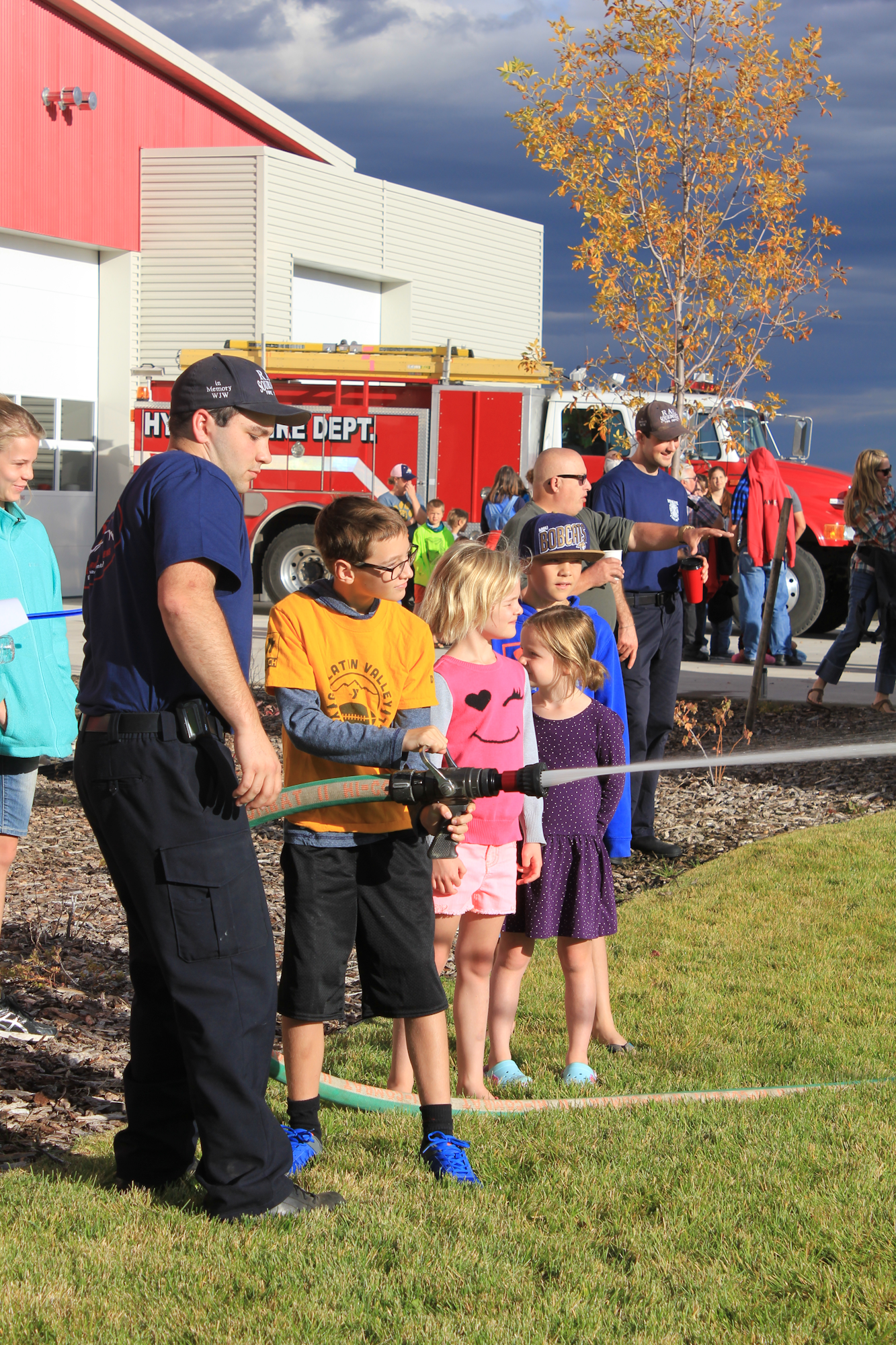 Kids learn to use a fire hose with a firefighter at a community event, with a fire truck in the background.