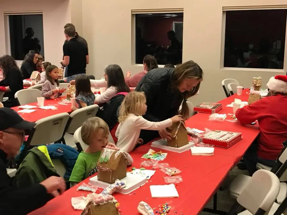 A group of children and adults decorate gingerbread houses at a festive gathering with red tablecloths and holiday treats.