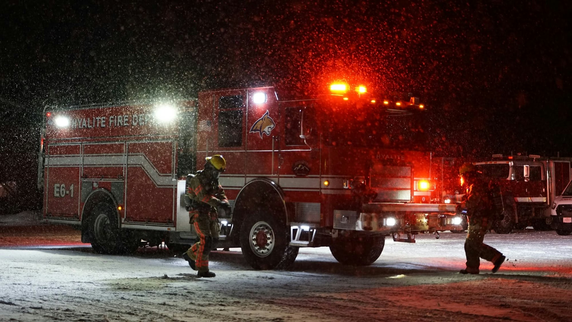 A snowy scene at night featuring a red fire truck with lights on and a firefighter preparing for action.