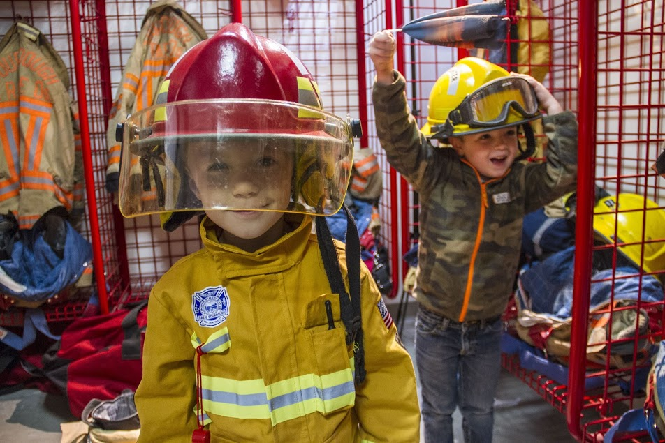 Two children dressed in fire gear, one smiling in a firefighter outfit and the other playfully holding a helmet. Fun and playful scene!