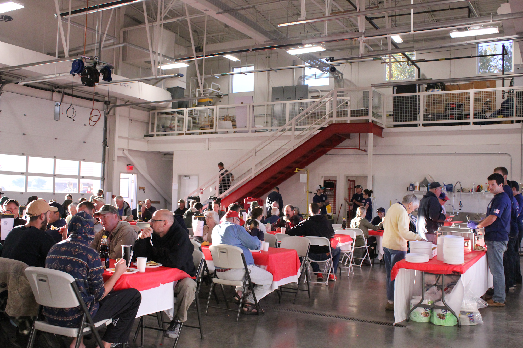 A large group of people dining inside a spacious hall, seated at tables with red tablecloths, enjoying a meal together.