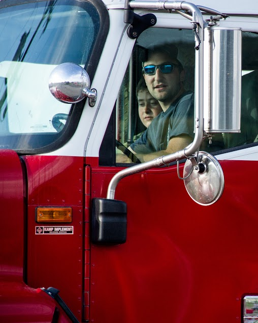A man and a woman are inside the cab of a red truck, both looking out the window. The man is wearing sunglasses.