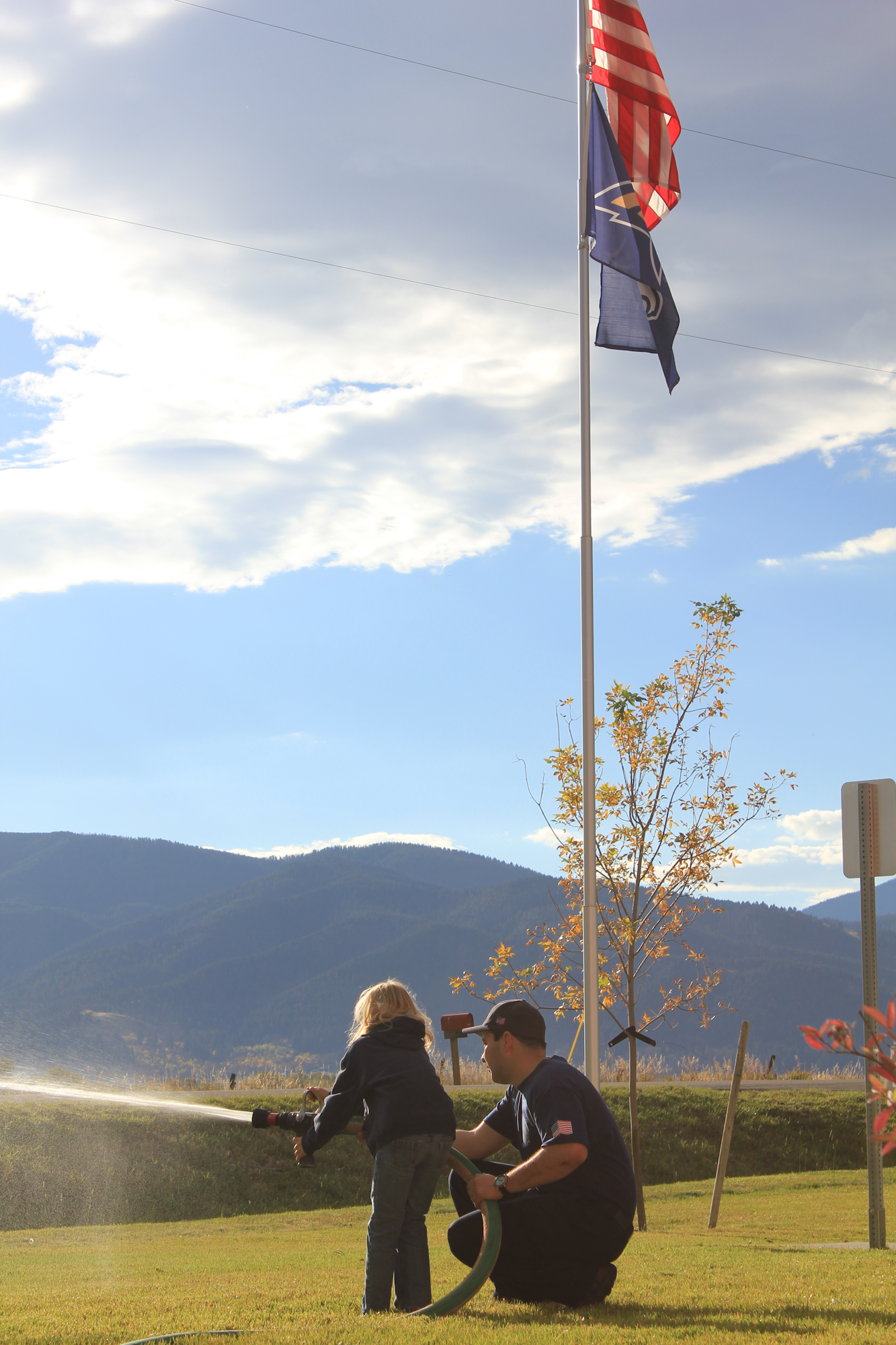 A child and an adult use a hose, with flags and mountains in the background, enjoying a sunny day outdoors.
