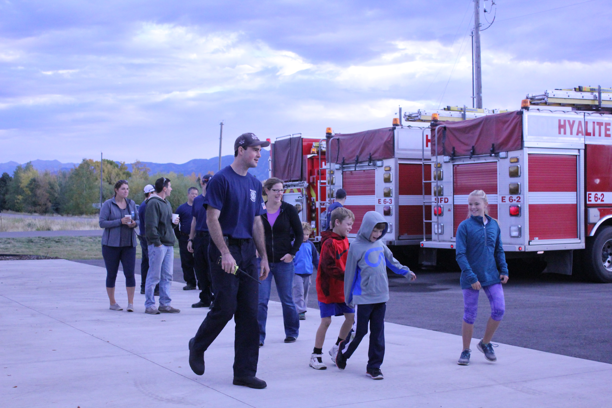 A group of people, including children, walks near fire trucks during an outdoor event. The setting is calm with a cloudy sky.