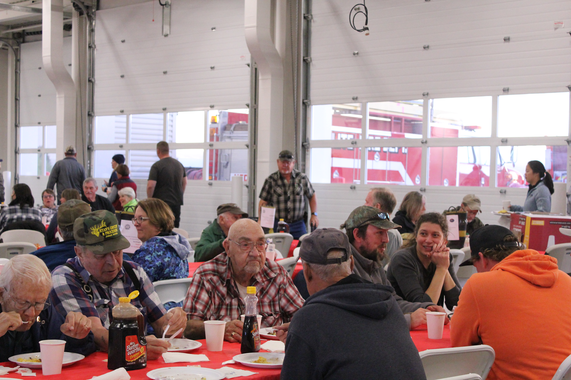 A group of people enjoying a meal together at tables with a red tablecloth in a spacious building.