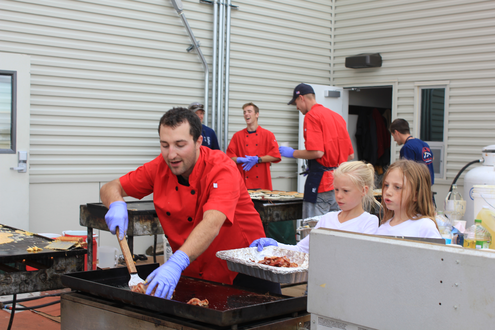 A group of cooks in red jackets prepares food outdoors, with children assisting and enjoying the cooking activity.