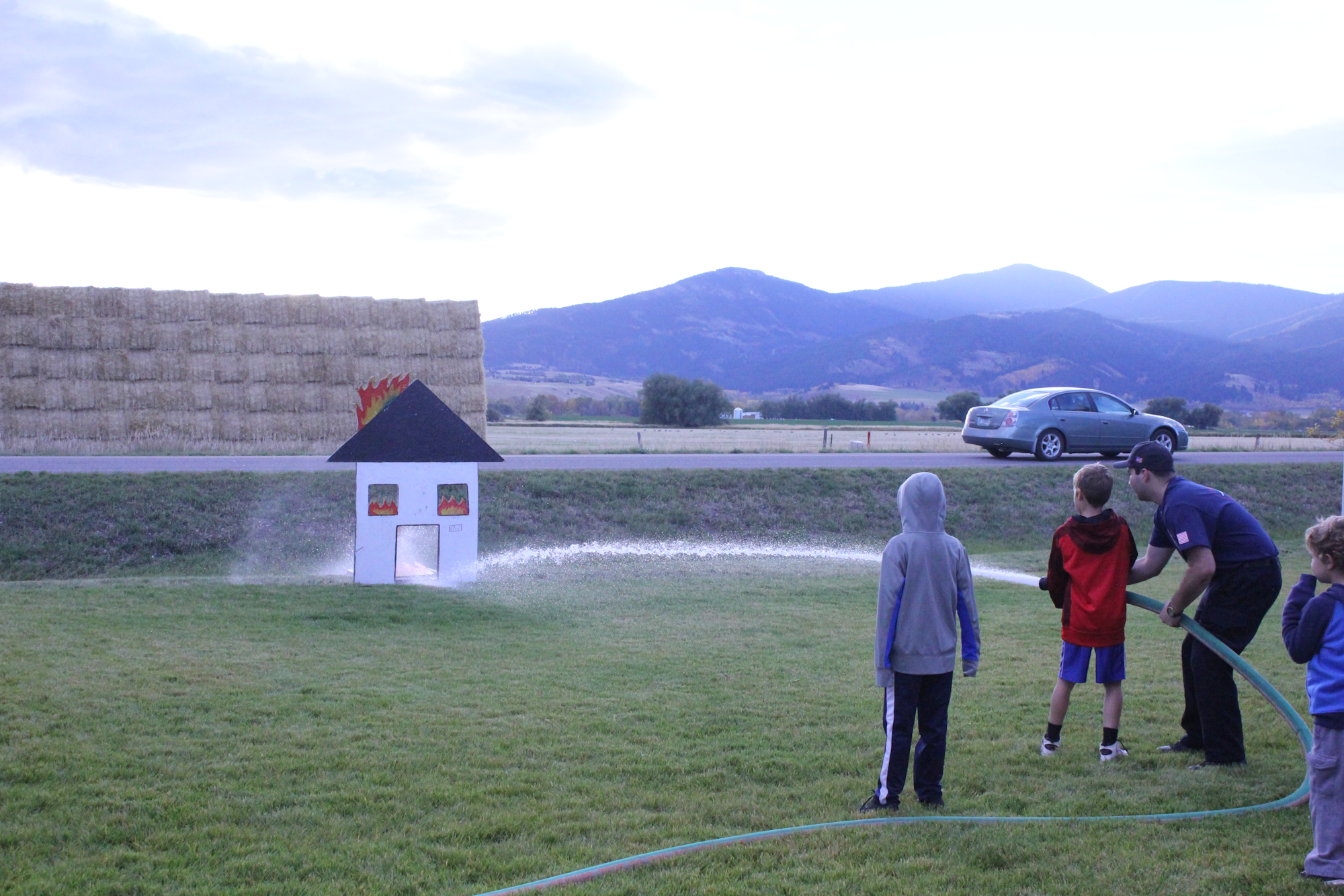 Kids and an adult practice firefighting techniques using a hose on a model house designed to look like it's on fire.