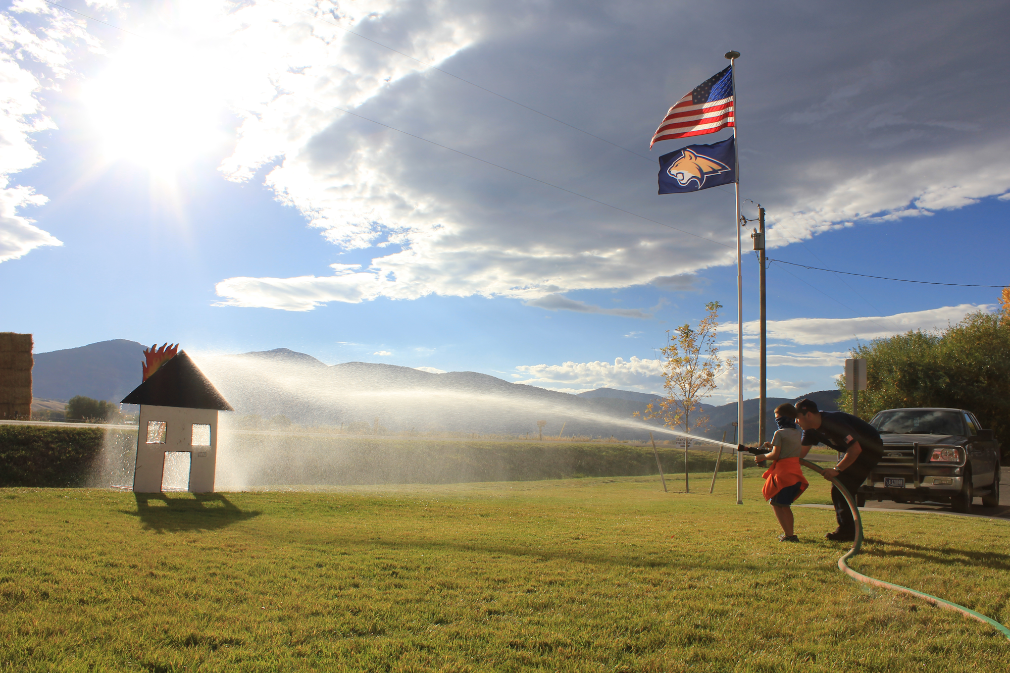 A person and a child play with a hose, spraying water near a toy house, with a clear sky and flags in the background.