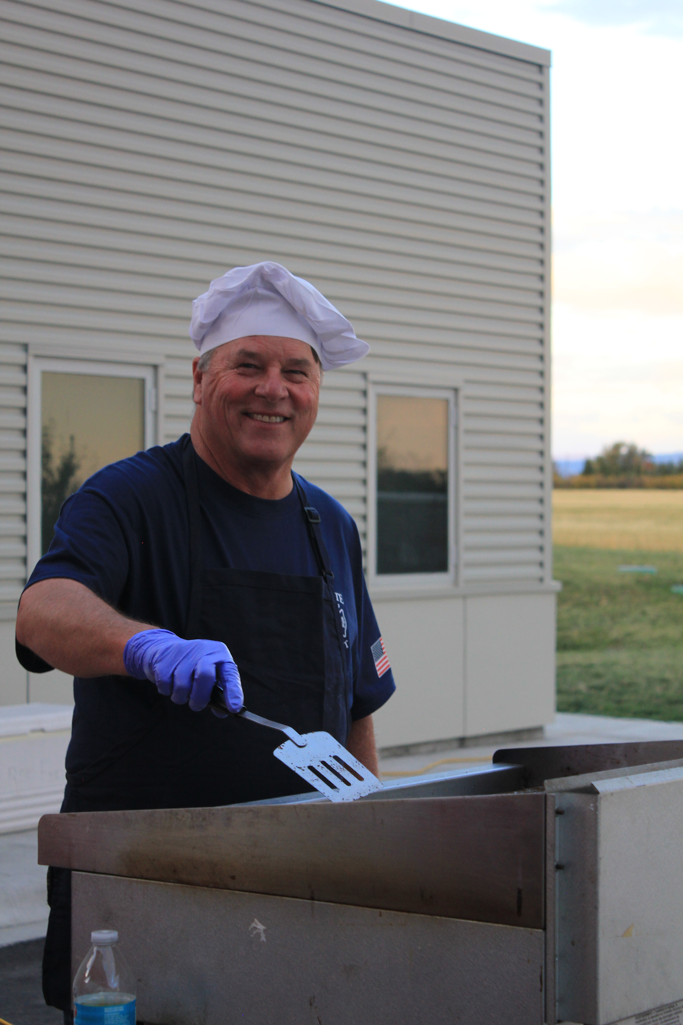 A smiling chef in a hat and gloves is cooking at a grill outdoors, with a blue sky and buildings in the background.