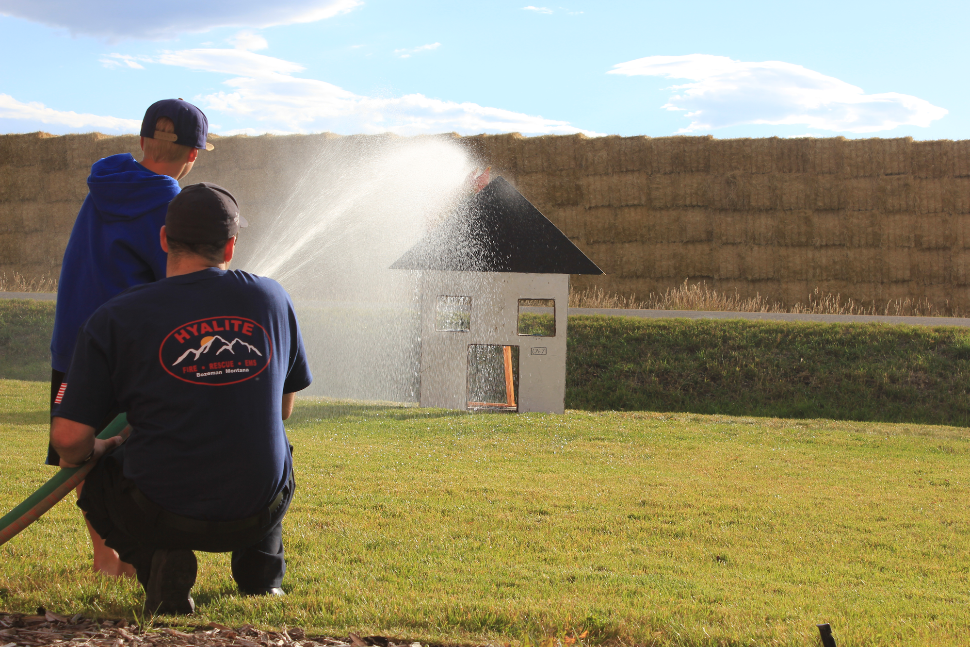 A person and a child spray water on a small model house from a hose, likely for fire safety training or practice.
