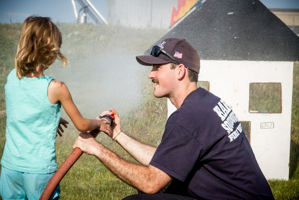 A child and a man are spraying water from a fire hose at a practice house, enjoying a fun and educational activity.