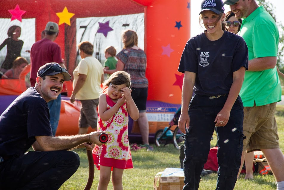 A fireman sprays water while a little girl smiles and covers her ears, enjoying a fun, festive outdoor event with families.
