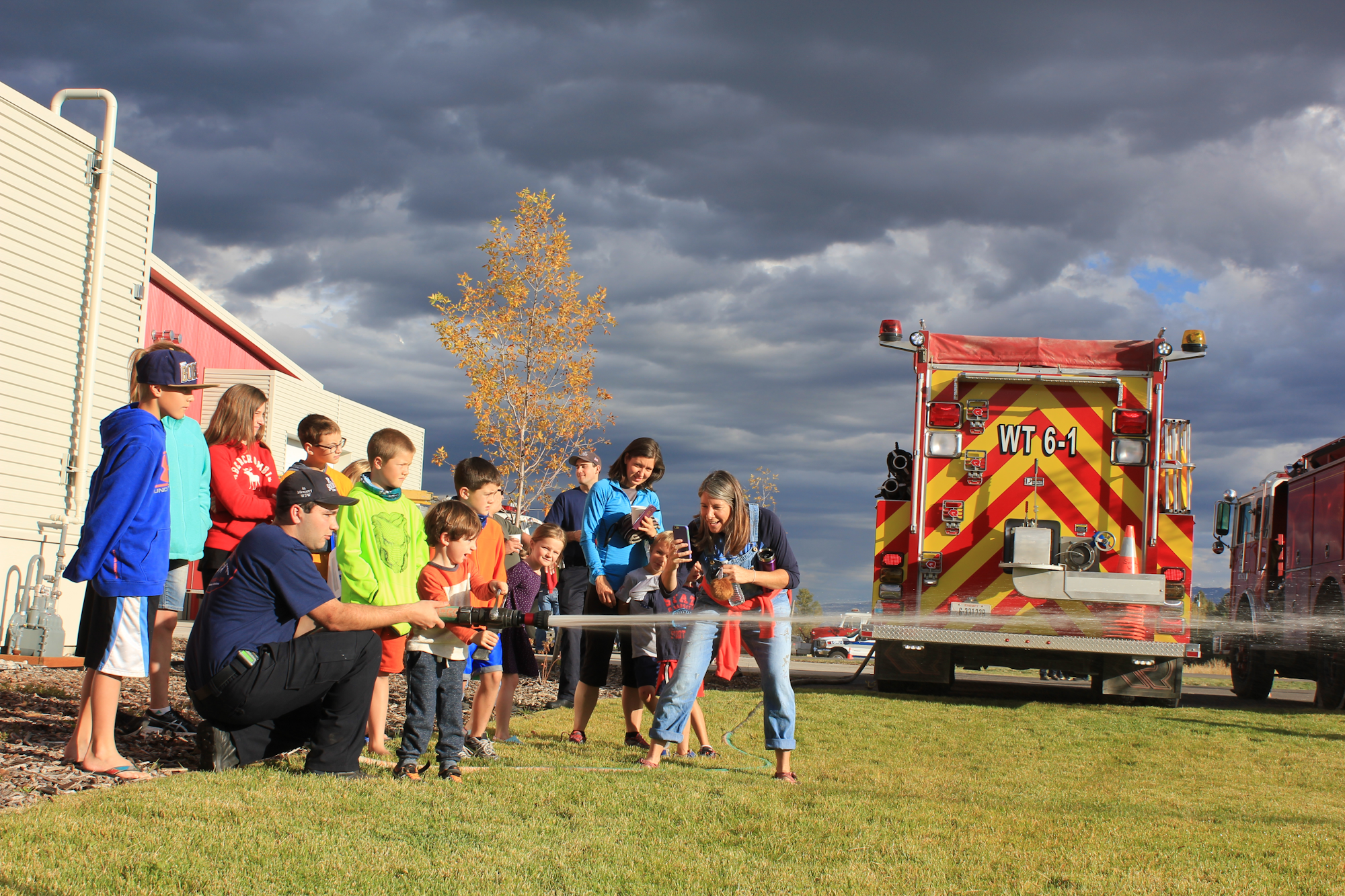A group of children and adults are participating in a fire safety demonstration near a fire truck, enjoying water spray and learning.