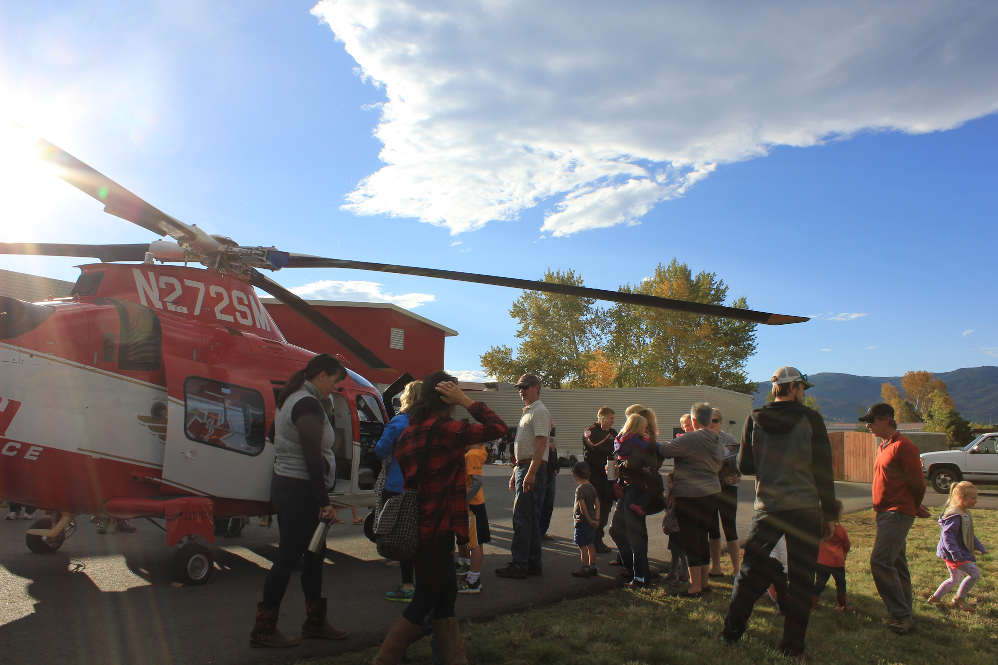 A crowd of people, including children, gathers around a red and white helicopter on a sunny day.