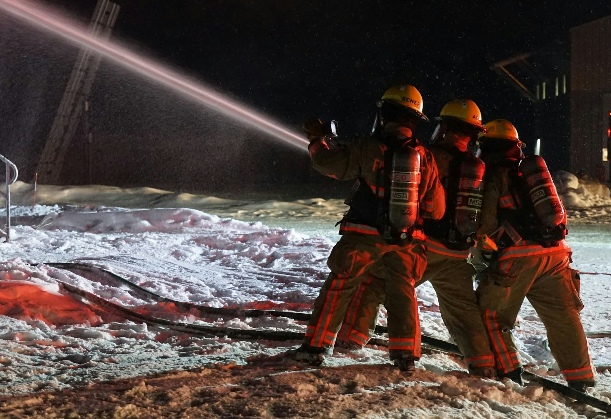 Firefighters in gear are spraying water on a snowy surface at night, focused on tackling a fire or emergency situation.