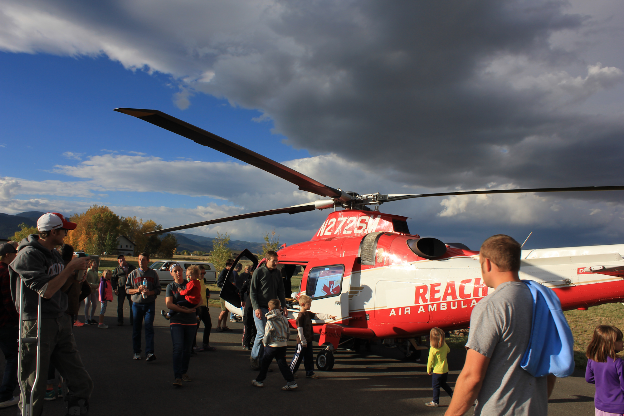 A crowd of people, including children, gathers around a red air ambulance helicopter under a cloudy sky.
