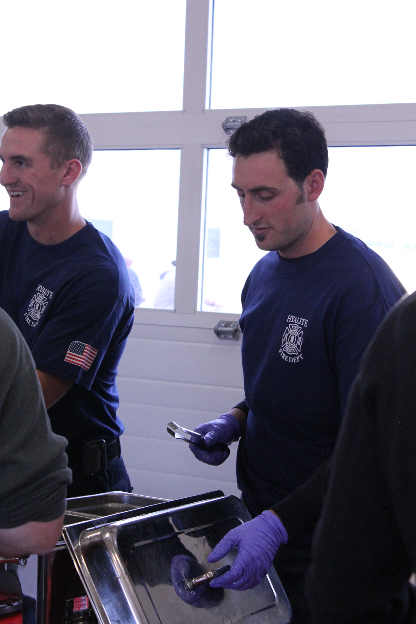 Two men in blue shirts, likely firefighters, prepare food in a kitchen setting. One is smiling while the other focuses on serving.