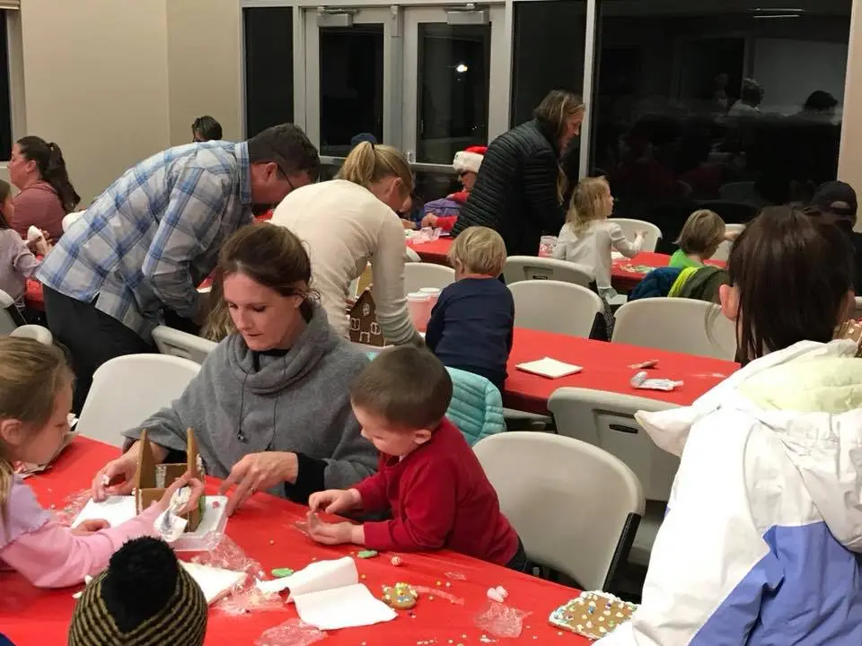 People, mostly families, gather around tables to create gingerbread houses at a festive event. Red tablecloths are visible.
