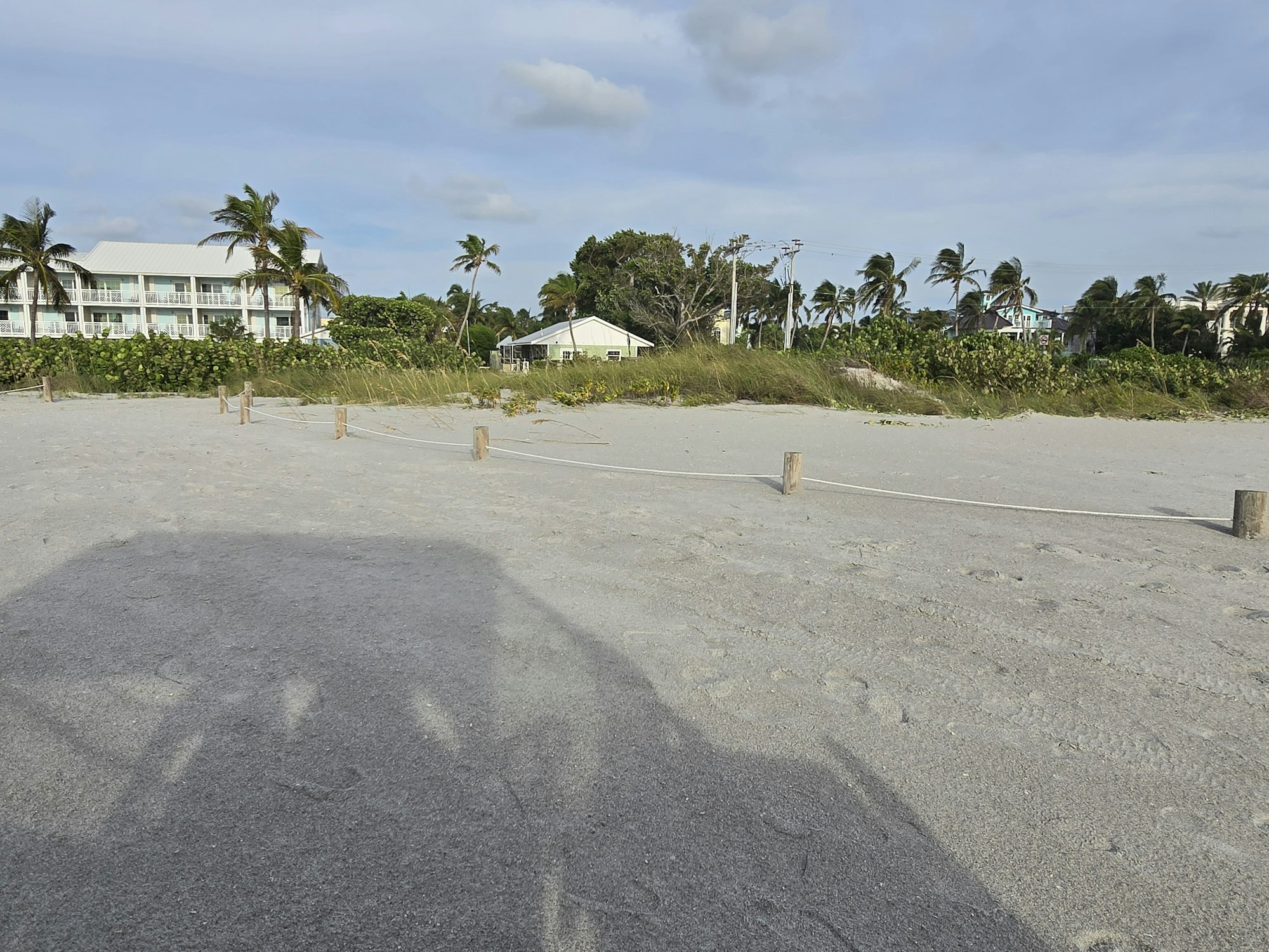 Beach parking area with sand, wooden posts, shadows, and buildings with palm trees in the background.
