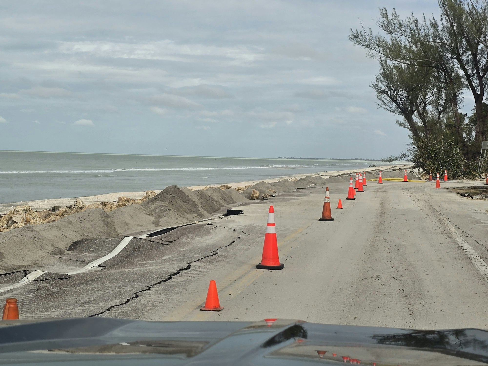 Damaged coastal road with traffic cones and sand piles along the sea.