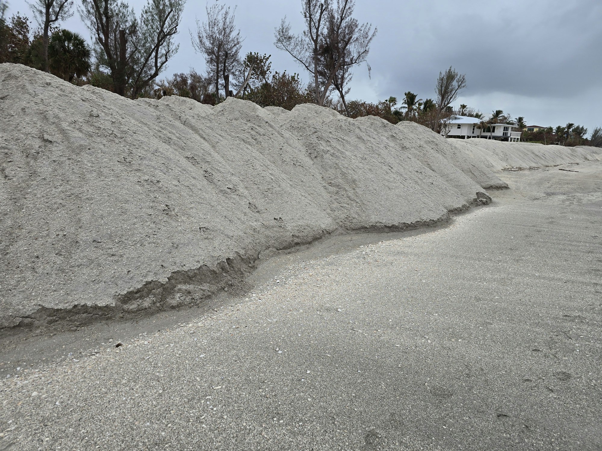 Piles of sand on a beach with trees and houses in the background under a cloudy sky.
