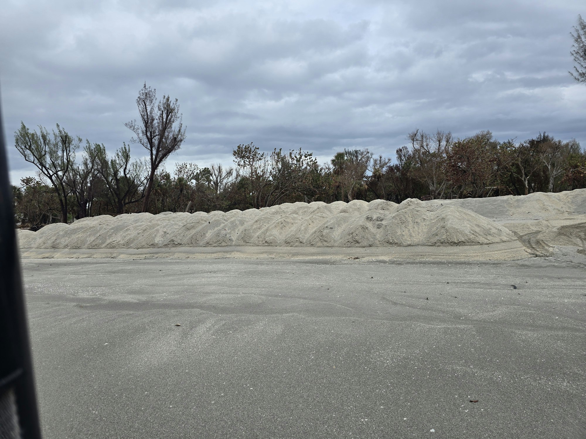Mounds of light-colored sand in an open area with trees and a cloudy sky in the background.