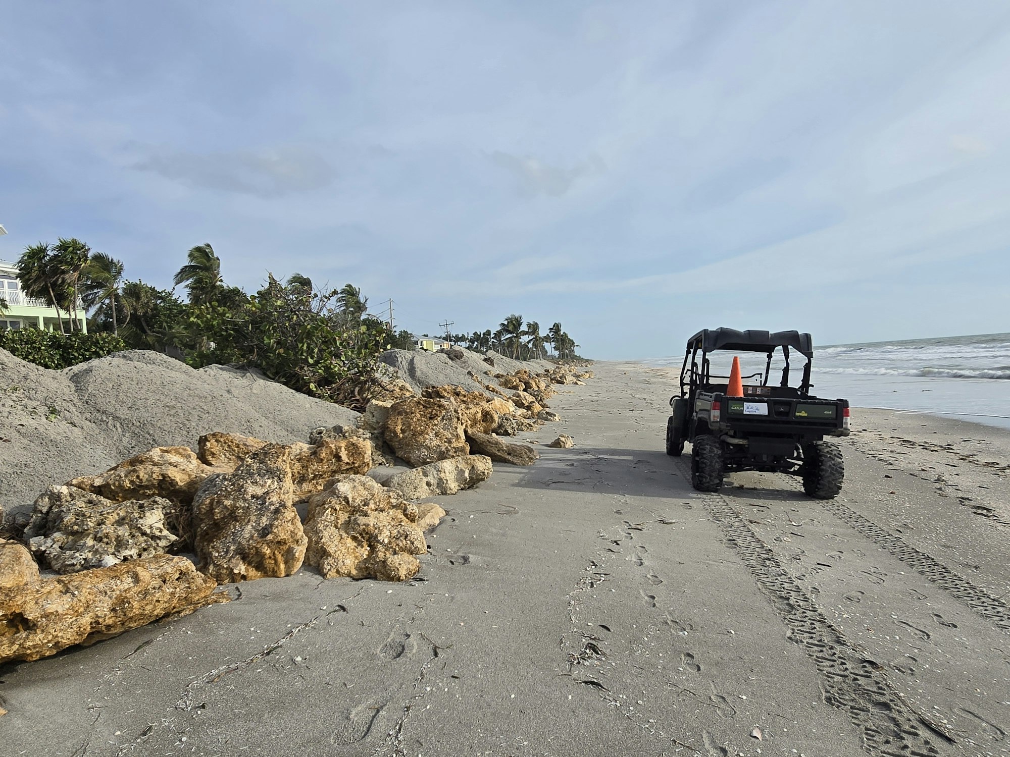 A utility vehicle on a beach with large rocks, footprints, and waves in the background under a partly cloudy sky.