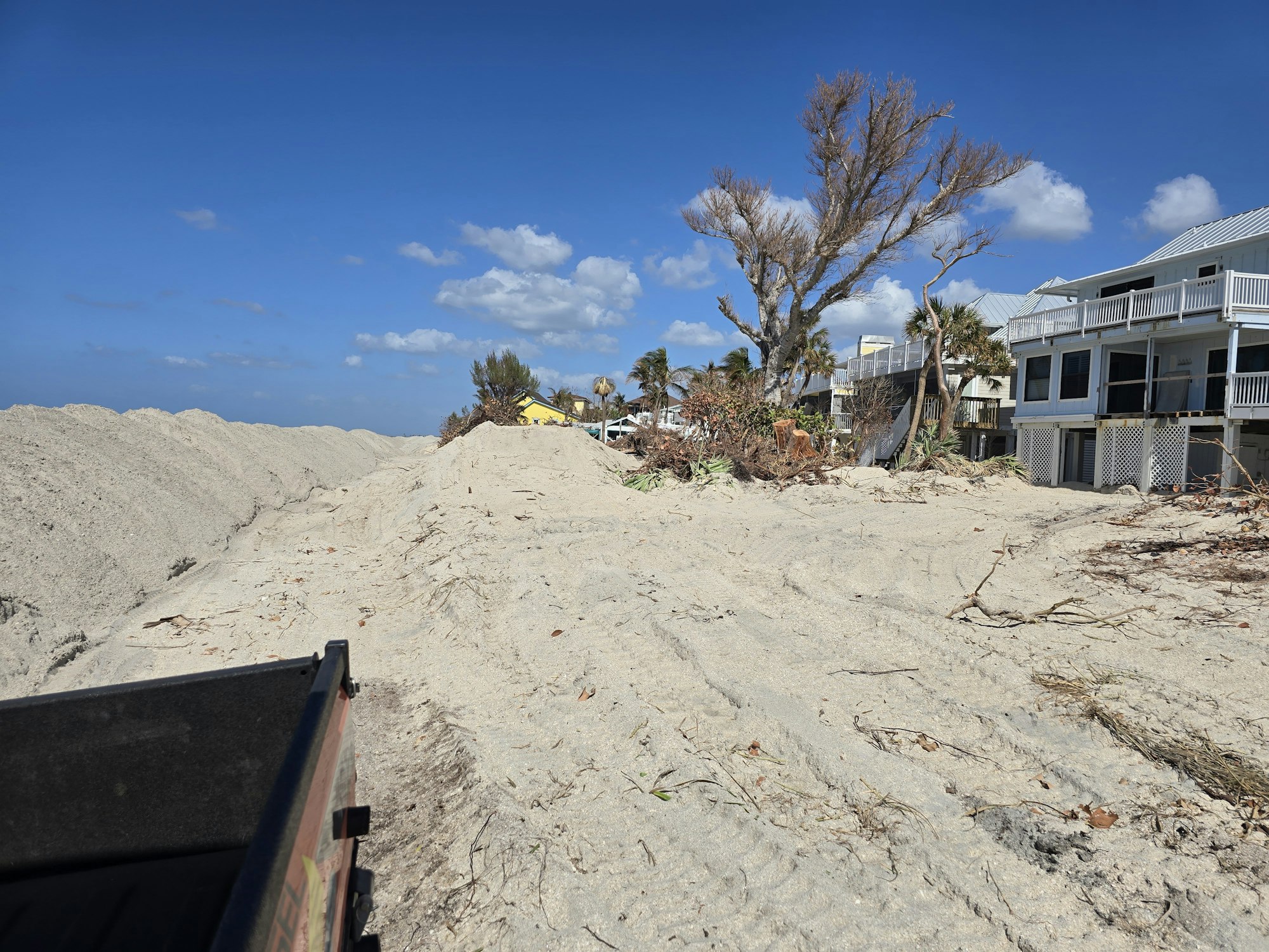 Sandy path with tall mounds, houses on the right, barren trees, and a clear blue sky.