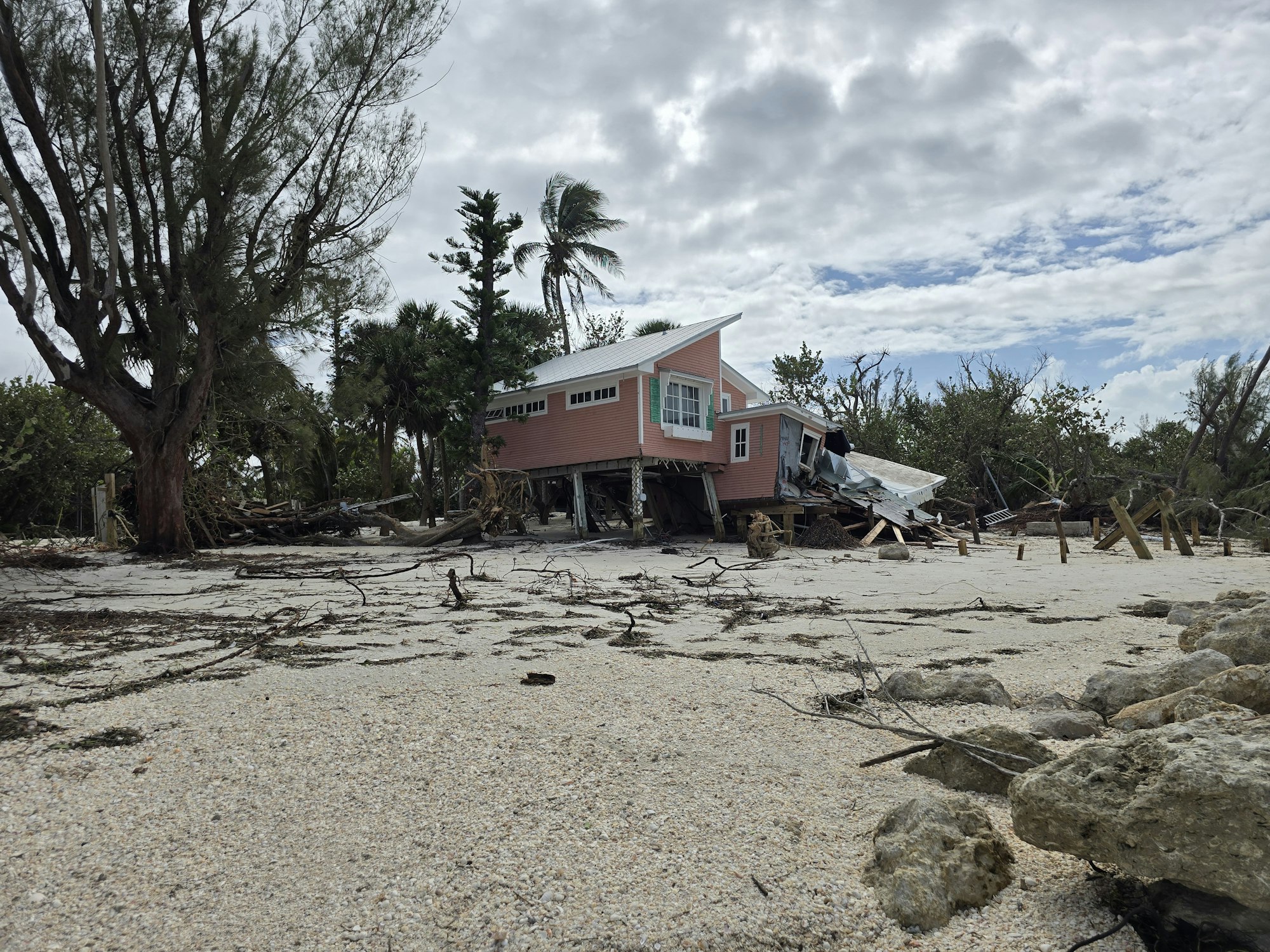 A damaged beach house surrounded by debris and fallen trees under a cloudy sky.