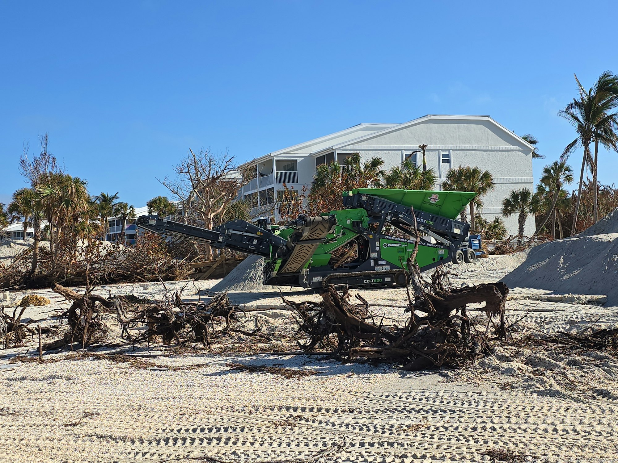 A green industrial machine on a sandy beach with uprooted trees and white buildings in the background.