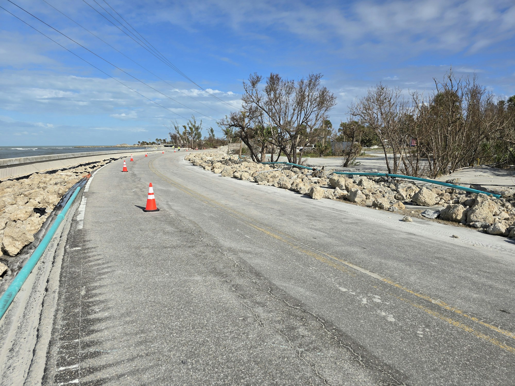 A road with traffic cones, debris, and damaged trees along the sides. Blue skies and a coastline in the background.