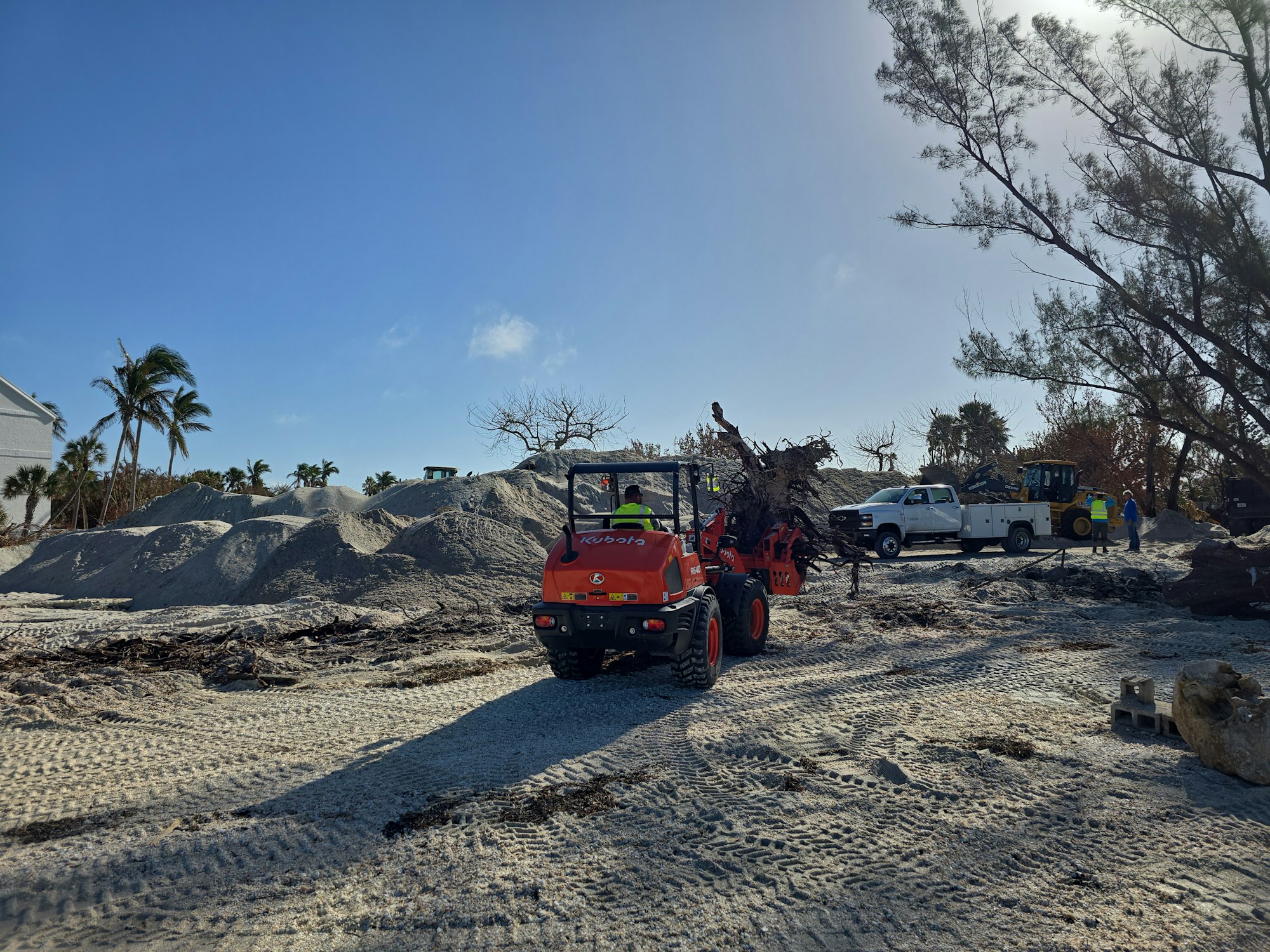 A construction site with a tractor, trucks, sand piles, and trees under a clear blue sky.